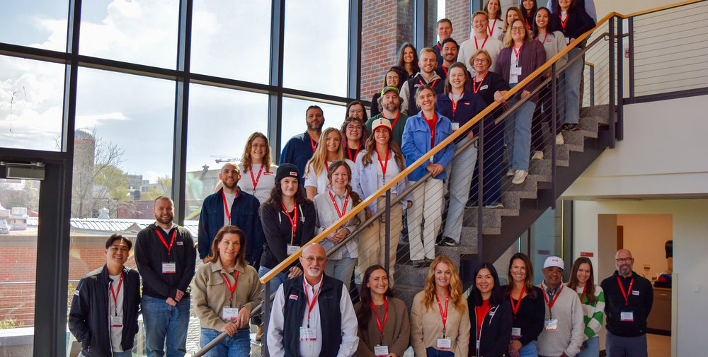 A group of people posing together on an indoor staircase in a modern building with large windows. Most are wearing matching red lanyards and badges.